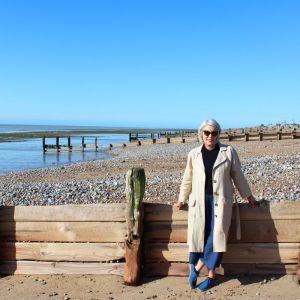 Fiona on the seafront at Worthing on a pebble beach, wearing her Elsie trousers and Lottie coat